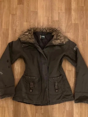 Mörkgrön vårjacka med fuskpäls - Snygg mörkgrön parkas från RAW med fuskpäls runt kragen. Perfekt till våren och skitsnygg, den har två fläckar på ena ärmen som visas i bilen men de går bort i tvätten. Den är också justerbar i midjan 