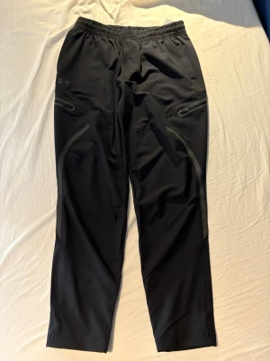 Svarta träningsbyxor Under Armour - Säljer ett par svarta träningsbyxor från Under Armour med elastisk midja och dragkedjeförsedda fickor. Byxorna har diskreta detaljer och är tillverkade i ett lätt, stretchigt funktionsmaterial som passar perfekt för träning eller löpning.