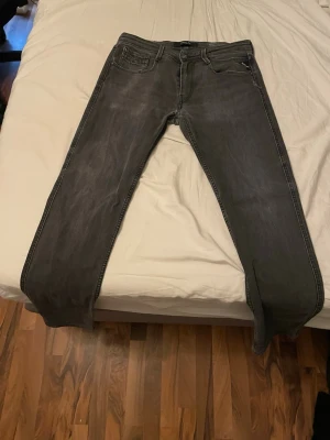 Replay anbas  - Säljer ett par gråa replay med raka ben och snygga kontrastsömmar. Jeansen har klassisk femficksdesign och detaljerade bakfickor med zigzag-mönster. Perfekta för en avslappnad och stilren look.