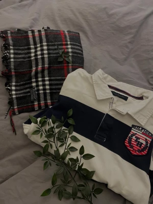 Randig rugbytröja från Tommy Hilfiger - Klassisk långärmad rugbytröja från Tommy Hilfiger i vitt och marinblått med breda ränder. Tröjan har en krage, knappar vid halsen och en stor broderad TH-logga i rött, vitt och blått på bröstet. Tillverkad i mjuk bomull för en bekväm känsla.