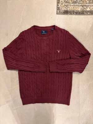 Vinröd kabelstickad tröja från GANT - Snygg vinröd kabelstickad tröja från GANT med rund halsringning och diskret logga broderad på bröstet. Tröjan har ribbade muddar vid ärmslut och nederkant. Perfekt för dig som gillar klassisk och stilren design.