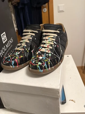 Maison Margiela Gats - Unika svarta sneakers med coola färgstänk i rött, blått, vitt, gult och grönt. Skorna har beige snörning och bruna sulor. Perfekt för dig som vill sticka ut med en kreativ och konstnärlig stil. Materialet är tyg och mocka. Con 9/10 inte speciellt mycket använda. Skicka dm för frågor/bilder. Box & dustbags ✅
