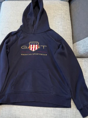 Mörkblå hoodie från Gant - Snygg mörkblå hoodie från Gant med broderad logga och texten 'American Sportswear' på bröstet. Klassisk känguruficka framtill och huva. Perfekt för en avslappnad och sportig stil.