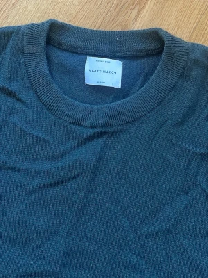Mörkgrön stickad tröja A Day's March - Mörkgrå stickad tröja från A Day's March i klassisk modell med rund halsringning och långa ärmar. Tröjan är tillverkad i mjuk merinoull och har ribbade muddar vid hals, ärmslut och nederkant. Storlek medium
