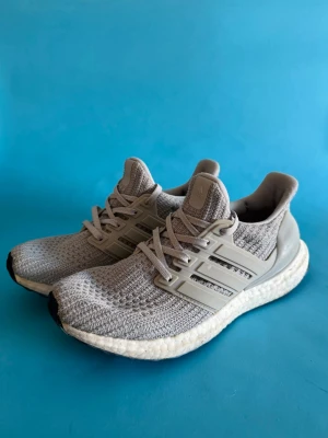 Grå Adidas Ultraboost sneakers - Snygga grå Adidas Ultraboost sneakers med stickad ovandel i mesh och klassiska tre ränder på sidorna. Skorna har en vit, dämpad Boost-sula och grå detaljer runt hälen. Perfekta för dig som gillar sportig och modern stil.