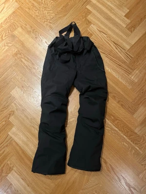 Svarta täckbyxor från Everest - Svarta täckbyxor från Everest med justerbara hängslen och dragkedjefickor. Perfekta för vinterns alla äventyr, tillverkade i slitstarkt och vattenavvisande material. Snygg och enkel design som funkar till det mesta.