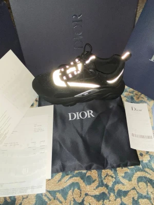 Svarta Dior sneakers med reflexdetaljer - Snygga svarta sneakers från Dior med feta reflexdetaljer som lyser upp i ljuset. Skorna har chunky sula, svarta skosnören och en modern, sportig design. Helt ny har aldrig använt.