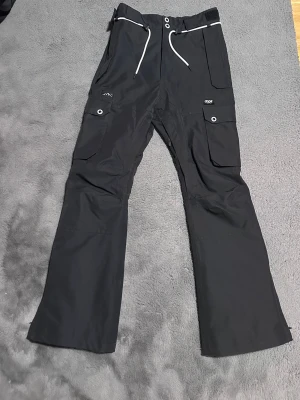 Svarta täckbyxor från Dope - Svarta täckbyxor från Dope med raka ben, stora cargofickor på sidorna och vita dragsnören i midjan. Byxorna har förstärkta knän och praktiska detaljer som tryckknappar och kardborre. Perfekta för vinterns äventyr och outdoor. Finns lite tecken på användning längst nere vid vaderna men inget man tänker på. Nypris 1999, mitt pris 799!