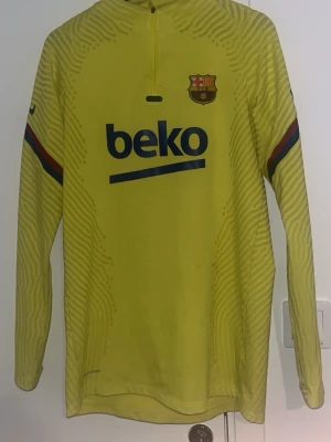 Gul långärmad FC Barcelona träningströja - Gul långärmad träningströja från Nike med FC Barcelona-logga och Beko sponsortryck på bröstet. Tröjan har halv dragkedja, subtila mönster i tyget och ränder i Barcelonas färger på ärmarna. Perfekt för fotbollsträning eller sportiga dagar.