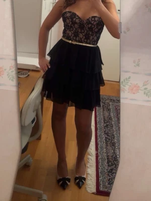 Svart spetsklänning med volanger - Snygg kort svart klänning med axelbandslös överdel i spets och volangkjol i flera lager. Klänningen har en beige underton upptill och ett smalt guldigt bälte i midjan. Perfekt för dig som vill sticka ut med en elegant look.