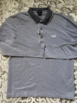 Grå långärmad piké från BOSS - Snygg grå långärmad piké från BOSS med klassisk krage och knappar framtill. T-shirten har en diskret logga på bröstet och är tillverkad i mjukt material som känns skönt mot huden. Perfekt för en stilren och avslappnad look. 