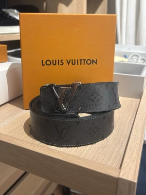 Svart Louis Vuitton monogram bälte - Snyggt svart bälte från Louis Vuitton med klassiskt monogrammönster och stor LV-logga i silver på spännet. Bältet är gjort i skinn och har en stilren, modern look som passar till många outfits.