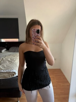 Svart axelbandslös topp med pärlor - Snygg svart axelbandslös topp med pärldetaljer som hänger längs nederkanten. Toppen har en figurnära passform och är perfekt för att skapa en cool look. toppen är croppad med bandeau-stil. Aldrig använd. Den öe H&M edition Nypris 500kr.