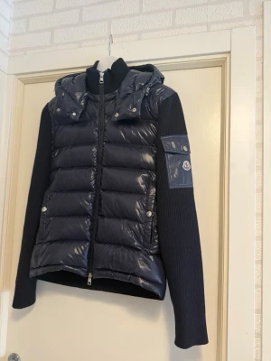 Navy cardigan från Moncler - Snygg navy cardigan från Moncler med glansig vadderad front och huva. Ärmarna är stickade i mjukt material och har en ficka med Moncler-logga på vänster arm. Jackan har dragkedja och tryckknappar framtill. Storlek M men passar bäst på S