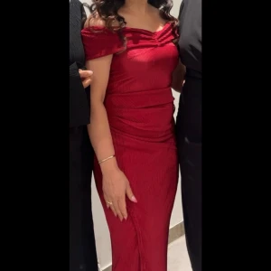 Röd klänning med slits - Säljer en elegant röd klänning med offshoulder och omlottdetaljer. Klänningen har en snygg slits på sidan, smickrande drapering och ribbad struktur. Jag har dock tagit bort bandet som knyts med kommer med ifall man vill sy den tillbaka. Använd en gång och är som ny.