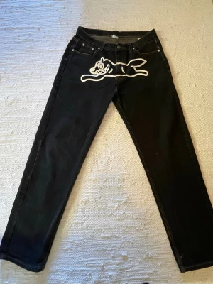 Svarta Billionaire Boys Club jeans - Unika svarta jeans från Billionaire Boys Club med vit kattgrafik framtill och astronauttryck på bakfickorna. Rak passform och klassisk femficksdesign. Tillverkade i bomull med cool streetwear-vibe. Någorlunda slitage runt fotleden. Förmodligen Replika.