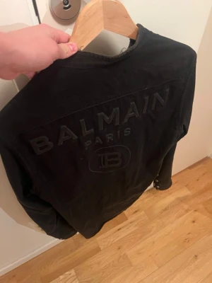 Balmain skjorta / jacka - Snygg svart jeansjacka från Balmain med stora broderade logotyper på ryggen och klassiska tryckknappar framtill. Jackan har två bröstfickor med lock och en stilren, rak passform. Perfekt för dig som vill ha en exklusiv streetwear-look.