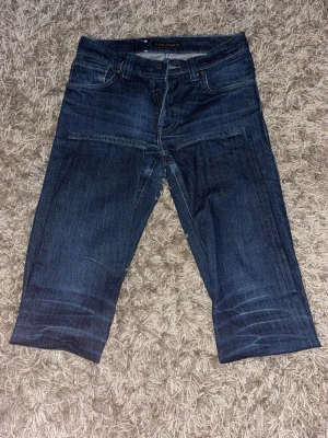 Mörkblå jeans från Nudie Jeans - Snygga mörkblå jeans från Nudie Jeans med klassisk femficksdesign och tydliga kontrastsömmar. Jeansen har raka ben och subtila slitningar framtill samt vågig brodyr på bakfickorna. Perfekta för en avslappnad och trendig look.
