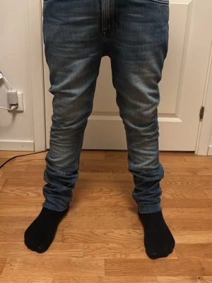 Blå slim fit jeans med låg gren - Säljer ett par blå slim fit jeans med låg gren och snygga slitningar. Jeansen har klassisk femficksdesign och är tillverkade i stretchigt denimtyg för extra komfort. Passformen är smal hela vägen ner och benen är något längre för en rynkad effekt vid anklarna.