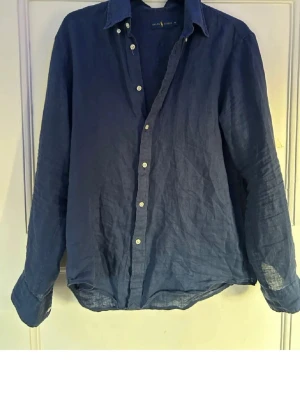 Annons till Simon!  - Snygg blå skjorta i linne från Ralph Lauren med klassisk krage och knappar framtill. Skjortan har lång ärm och en avslappnad passform, perfekt för en stilren look. Märkeslogga på insidan av kragen.