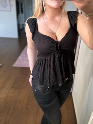 Svart topp med volang och knyt - Säljer en svart topp med volangkant och knyt framtill. Toppen har djup urringning och smockad struktur i tyget. Perfekt att styla med jeans eller kjol💗
