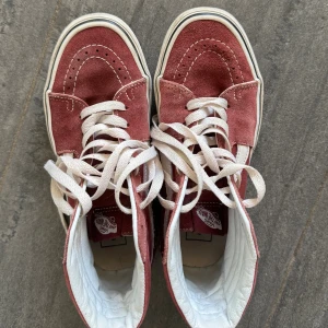Vinröda Vans  - Säljer ett par vinröda Vans sneakers i mocka med vita detaljer och klassisk vit sula. Skorna har högt skaft, vita skosnören och den ikoniska Vans-sidan i vitt. Perfekta för dig som gillar streetwear och vill ha en retro look.