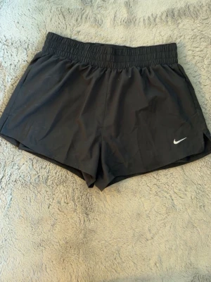 Svarta träningsshorts från Nike - Svarta träningsshorts från Nike med elastisk midja och klassisk vit logga på ena benet. Lätta och bekväma shorts i syntetmaterial, perfekta för sport eller gym. Modellen är loose fit och har en enkel, stilren design.