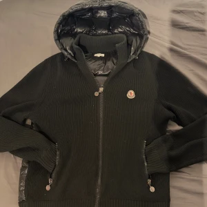 Svart stickad jacka från Moncler - Säljer en svart stickad jacka från Moncler med quiltad huva och innerfoder. Jackan har dragkedja framtill, två fickor med dragkedja och Moncler-logga på bröstet. Perfekt för dig som vill ha en snygg och bekväm jacka med exklusiv känsla, från tidigt 2010 tal med certi logo ingen Nfc eftersom att dehär var innan dess tid, lite hål i armhålan men går enkelt o laga dessutom märker man knappt av de!