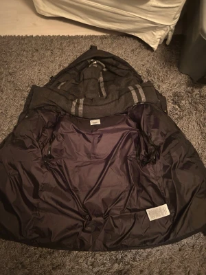 Mörkgrå Burberry kappa med huva - Säljer en snygg mörkgrå kappa från Burberry med huva och klassiska ränder på insidan. Kappan har ett mjukt foder och är perfekt för kyliga dagar. Materialet är ullblandning med detaljer i syntet. Stilsäker och tidlös design.