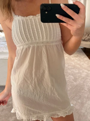 Vit kort sommarklänning med volang - Söt vit kort klänning med smala axelband och volangkant nertill. Överdelen har broderade detaljer och rynk, medan kjolen är luftig och lätt. Perfekt för varma dagar och har en romantisk känsla.