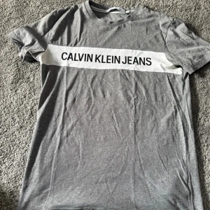  Calvin Klein t-shirt - Snygg grå t-shirt från Calvin Klein Jeans med vit panel och stor logga över bröstet. Klassisk passform och rund halsringning. Perfekt till jeans eller shorts för en clean och stilren look.