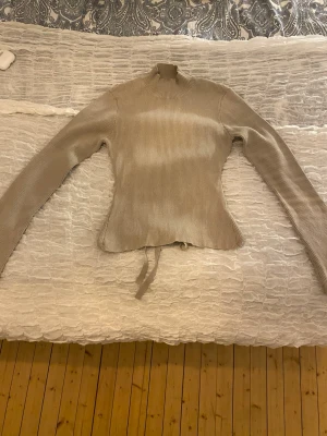 Beige ribbad topp med knytband - Trendig beige ribbad topp med lång ärm och hög hals. Toppen har figurnära passform och snygga knytband i midjan som ger en unik detalj. Endast använd fåtal gånger, köpt på vinted ursprungligen från pretty lavish 💕