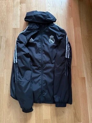 Svart Real Madrid vindjacka Adidas - Svart vindjacka från Adidas med Real Madrid-logga på bröstet och vita ränder längs ärmarna. Jackan har huva, dragkedja framtill och elastiska muddar vid ärmsluten. Perfekt för dig som gillar sportig stil och vill visa ditt lagstöd. Pris går att diskutera.