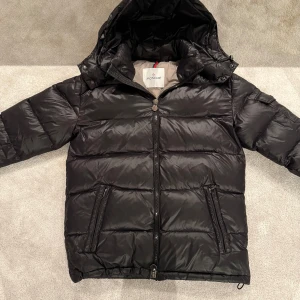 Svart Moncler pufferjacka med huva - Säljer en svart pufferjacka från Moncler med huva och dragkedja framtill. Jackan har två snedställda fickor med dragkedja och en ärmficka med Moncler-logga. Klassisk quiltad design och riktigt skön passform för kalla dagar.