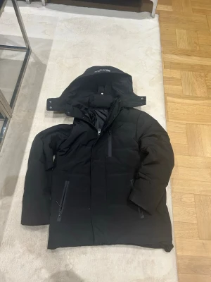 Svart dunjacka med huva S  - Säljer en svart dunjacka med avtagbar huva och dragkedjor på fickorna. Jackan har en stilren design och är perfekt för kalla vinterdagar. Fodrad för extra värme och har en hög krage som skyddar mot vind. Passar dig som vill ha en enkel men snygg vinterjacka.