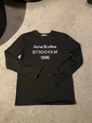 Svart long sleeve Acne Studios - Svart långärmad t-shirt från Acne Studios med vitt tryck framtill där det står 'Acne Studios STOCKHOLM 1996'. Klassisk rund hals och relaxed fit. Perfekt för dig som gillar stilrena och trendiga plagg med tydlig logga.