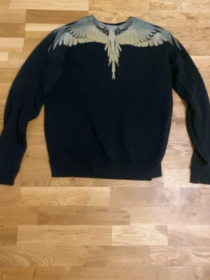 Svart sweatshirt Marcelo Burlon - Svart sweatshirt från Marcelo Burlon County of Milan med ikoniskt vingtryck i grått och beige över axlar och halsringning. Tröjan har rund hals och ribbade muddar. Perfekt för dig som gillar streetwear och unika detaljer.