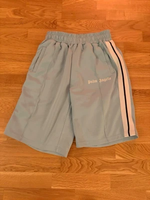 Palm Angels ljusblå shorts med revär - Palm Angels shorts i ljusblått med vita och svarta revärer längs sidorna. Elastisk midja och två fickor framtill. Ikonisk logga tryckt på vänster ben. Perfekta för en avslappnad streetwear-look. pris kan diskuteras!!