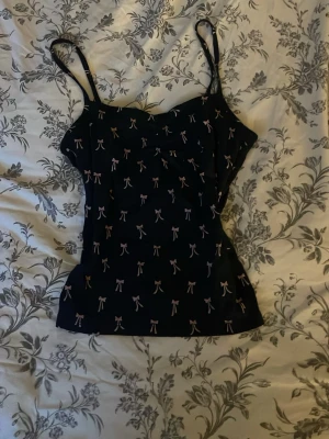 Svart linnetopp med rosettprint - Söt svart linnetopp med tunna axelband och små rosa rosetter som mönster. Toppen har en enkel, rak passform och är perfekt för en avslappnad look. Materialet känns mjukt och lätt mot huden.