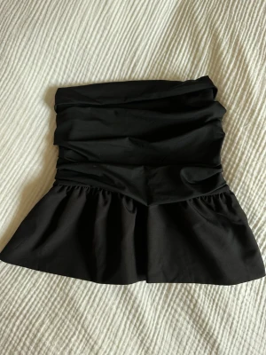 Svart bandeau tubtopp med volang - Svart bandeau tubtopp med volangkant nertill. Toppen har en snygg draperad effekt upptill. Aldrig använd 