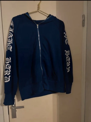 Blå zip hoodie med tryck på ärmar - Mörkblå hoodie med dragkedja framtill och vit text i gotisk stil längs båda ärmarna. Hoodien har huva och två fickor framtill. Materialet ser ut att vara mjuk bomullsblandning, perfekt för en avslappnad streetwear-look.