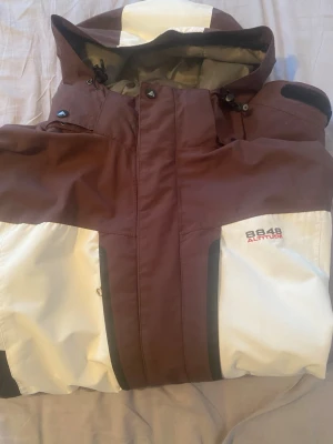 8848 Altitude brun och vit vindjacka - Snygg vindjacka från 8848 Altitude i brunt och vitt med svarta detaljer. Jackan har huva med dragsko, två framfickor med dragkedja och logga på bröstet. Perfekt för blåsiga dagar och outdoor-aktiviteter. Storlek M+L