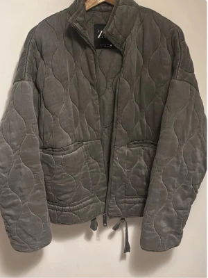 Grå quiltad jacka från Zara - Snygg grå quiltad jacka från Zara med vågigt mönster och två stora fickor framtill. Kommer utan banden eftersom jag tog av den! Färgen e typ grå/grön 