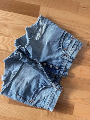 Levi's 501 ljusblå jeansshorts - Säljer ett par klassiska Levi's 501 jeansshorts i ljusblå denim. Shortsen har råa, fransiga kanter och slitna detaljer framtill för en avslappnad look. Knappar i gylfen och femficksmodell. Perfekta för sommaren!