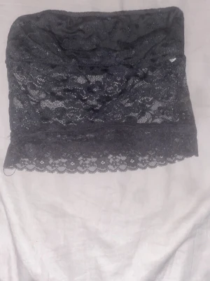 Svart spetsbustier bandeau - Säljer en svart bandeau bustier i mjuk spets med blommigt mönster. Toppen är axelbandslös och har en elastisk kant upptill för bättre passform. Perfekt att bära under en skjorta eller som den är. Materialet är stretchigt och bekvämt.