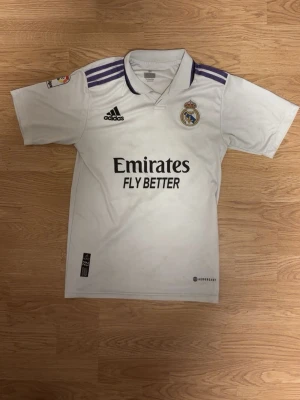 Real Madrid matchtröja Adidas vit - Säljer en vit Real Madrid matchtröja från Adidas med klubbmärke på bröstet och svart Adidas-logga. Tröjan har lila detaljer på axlarna, svart text 'Emirates Fly Better' och klassisk krage. Perfekt för fotbollsfans som vill visa sitt stöd på planen eller läktaren.