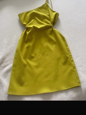 Gul one shoulder klänning - Trendig gul klänning med one shoulder-design och smal axelrem. Klänningen har en snygg cut-out detalj i midjan och är kort i modellen. Perfekt för dig som vill sticka ut med en färgstark look.