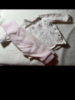 Sött babyset med kattmotiv från H&M - Ett gulligt babyset från H&M i storlek 56, med en långärmad vit body i ekologisk bomull och rosa kattmotiv samt knappar på sidan. Tillhörande ljusrosa mjuka byxor med breda muddar. Perfekt för små bebisar som vill vara både bekväma och stiliga.