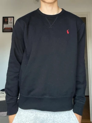 Svart sweatshirt från Polo Ralph Lauren - Klassisk svart sweatshirt från Polo Ralph Lauren med rund halsringning och röd broderad logga på bröstet. Tröjan har ribbade muddar vid ärmslut och nederkant, och är tillverkad i mjuk bomullsblandning. Perfekt för en clean och stilren look.