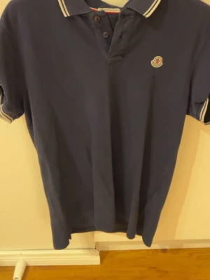 Marinblå pikétröja från Moncler - Snygg marinblå pikétröja från Moncler med vitrandiga detaljer på krage och ärmslut. Klassisk polokrage med knappar och Moncler-logga på bröstet. Perfekt till jeans eller shorts för en clean look.